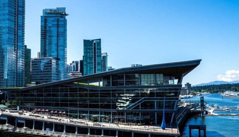 Modern architectural structure with a distinctive roof design, located near water and surrounded by tall buildings.